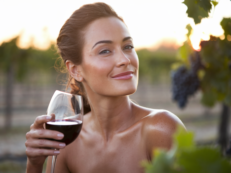 Mulher com pele saudável apreciando vinho tinto em um vinhedo ao pôr do sol, representando o conceito de que vinho faz bem para a pele.