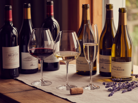 Três taças com vinhos bons e baratos até R$ 20 sobre uma mesa rústica, com garrafas ao fundo.